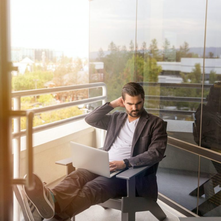 man sitting on gray arm chair using silver laptop computer on building balcony at daytime