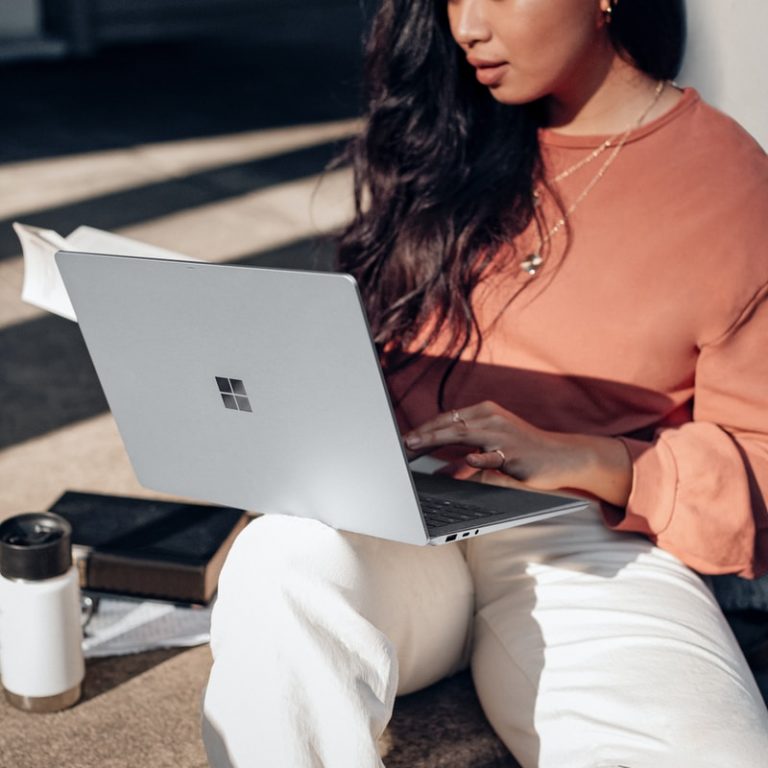 woman in orange long sleeve shirt and white pants sitting on floor using Surface Laptop
